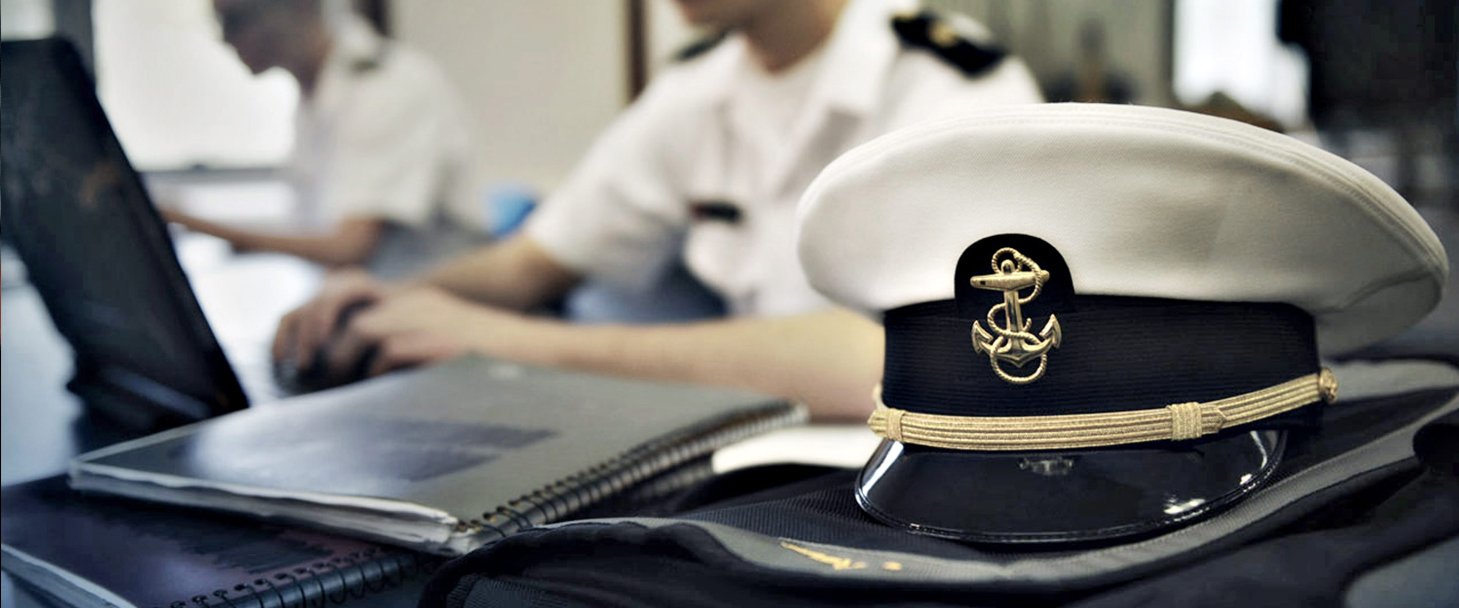 A U.S. Navy Officer's cap sits on a table with notebooks in the foreground while Sailors participate in an undergraduate college class.