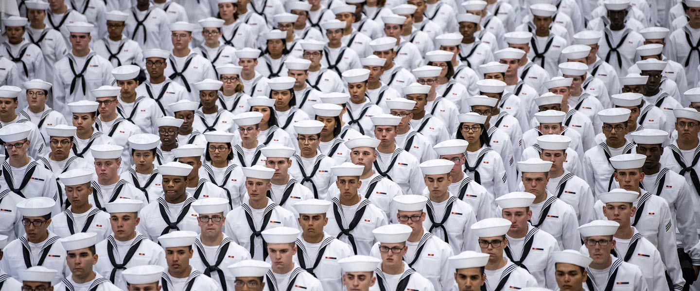 A large group of U.S. Navy recruits stand in group formations wearing formal attire