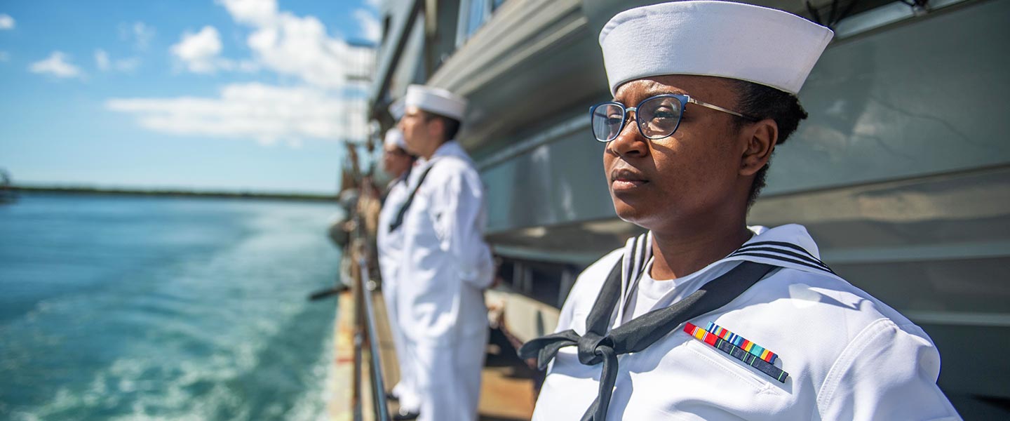 A diverse group of Navy sailors in formal sailor uniform stand at attention on the deck of a Navy aircraft carrier