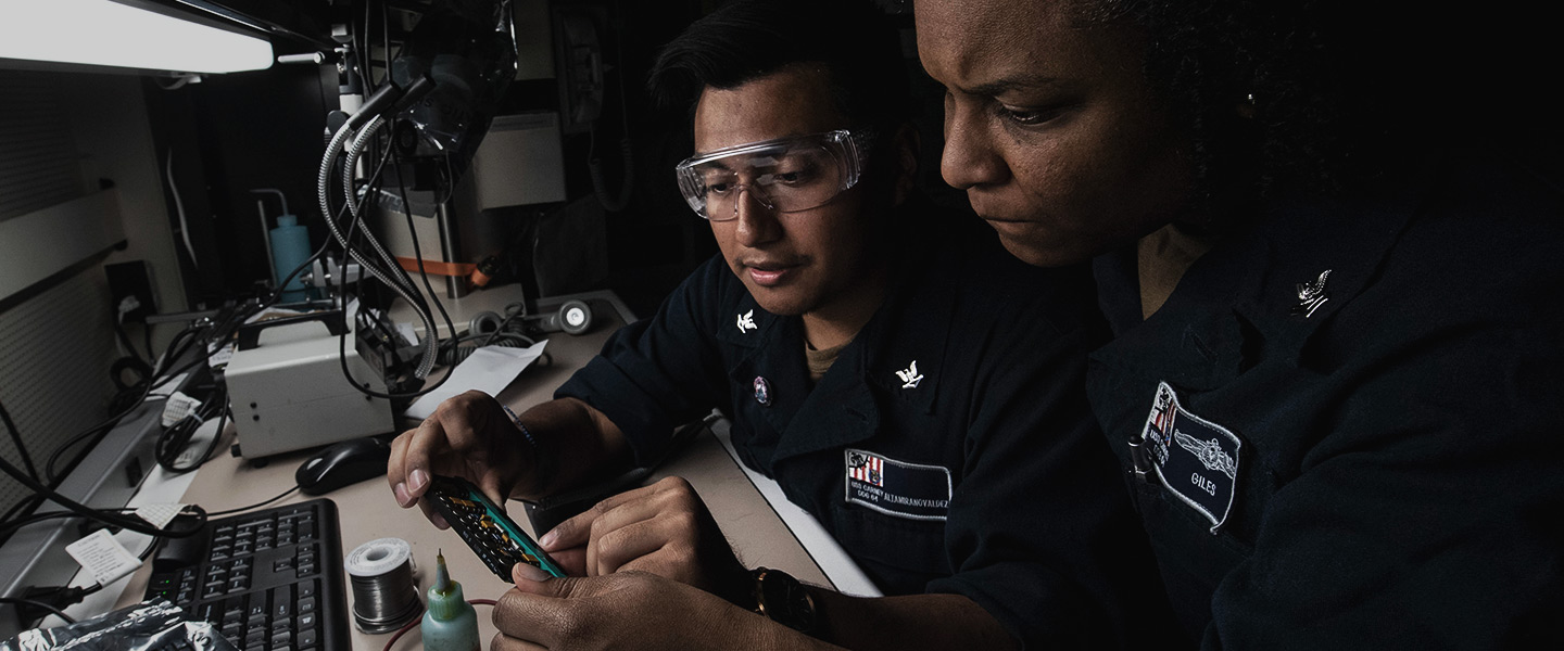 Two United States Navy Electronics Technicians assess a circuit board prior to soldering while conducting routine maintenance checks aboard the USS Carney.