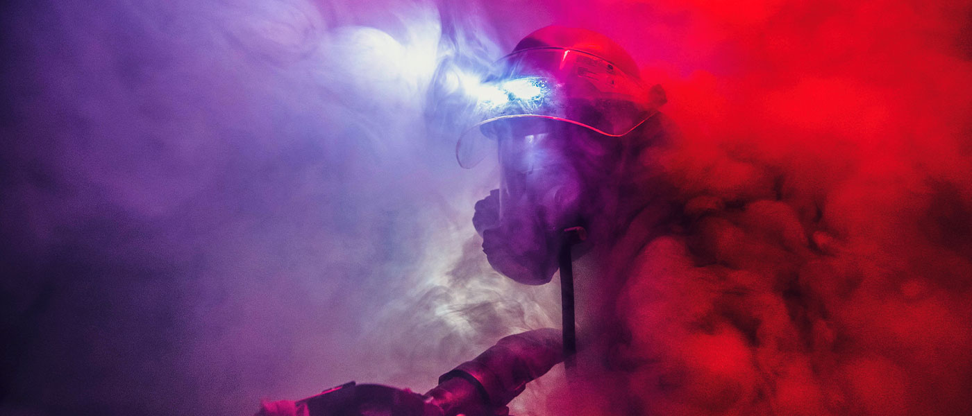 A Navy first responder in firefighter gear moves cautiously through a room full of smoke