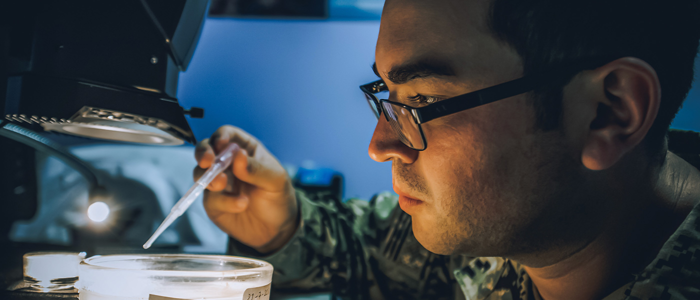 A United States Navy Sailor uses a high-powered microscope to analyze a test sample while working in a laboratory aboard a ship.