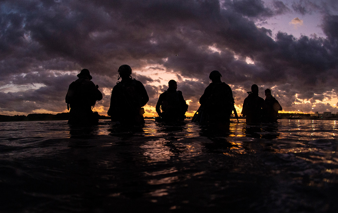 Sailors from a Naval Special Warfare Group conduct dive operations at sunset, silhouetted against dramatic clouds and reflective waters.