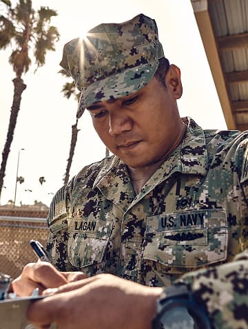 A U.S. Navy Sailor is filling out a form while in uniform with the sun reflecting behind him. 