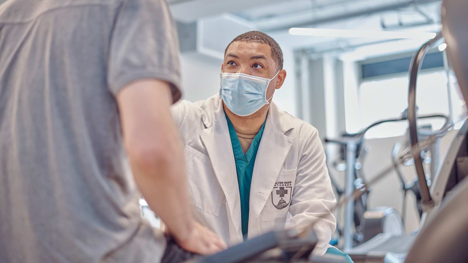 U.S. Navy Hospital Corpsman Dominique Velazquez wears a white medical coat and protective face mask while checking on a fellow navy sailor patient