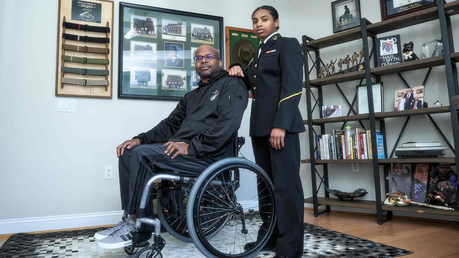 US Naval Academy Midshipman Kaylah Gillums stands in uniform behind her father, who is seated in his wheelchair.