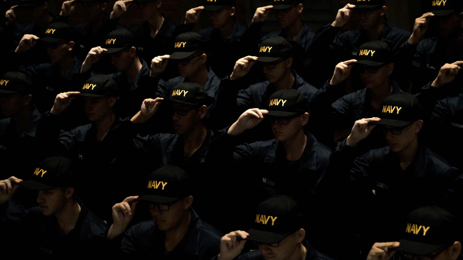 A large group of United States Navy sailors gather at a ceremony and salute together while wearing Navy baseball caps