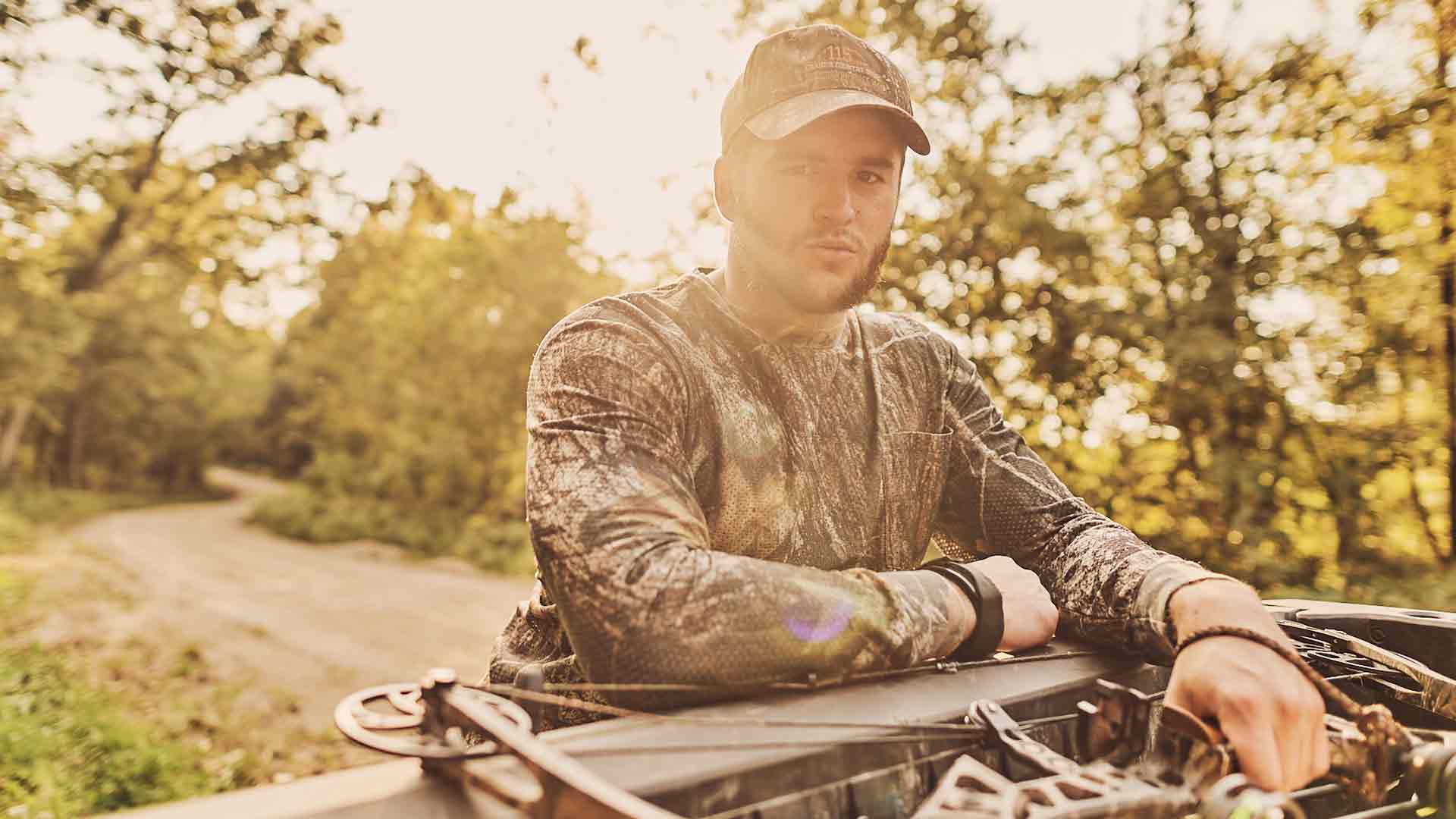 Colten Hubbard poses for a photo on a dirt road near the wilderness wearing camo hunting gear and holding a hunting bow