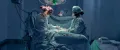 Navy surgeons in a brightly lit operating room, one wearing a patterned cap, performing surgery with a third surgeon observing in the background. 