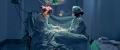 Navy surgeons in a brightly lit operating room, one wearing a patterned cap, performing surgery with a third surgeon observing in the background. 