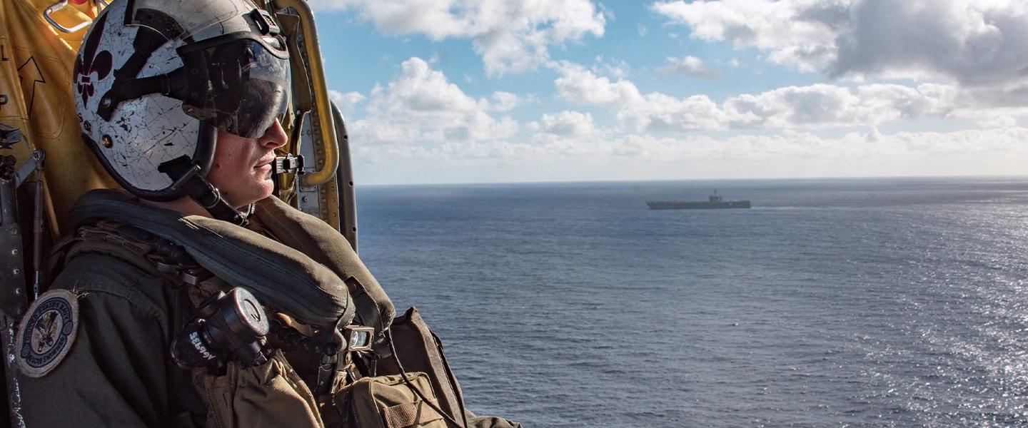 A U.S. Navy Sailor looks out over the ocean from inside a helicopter as an aircraft carrier transits the sea.