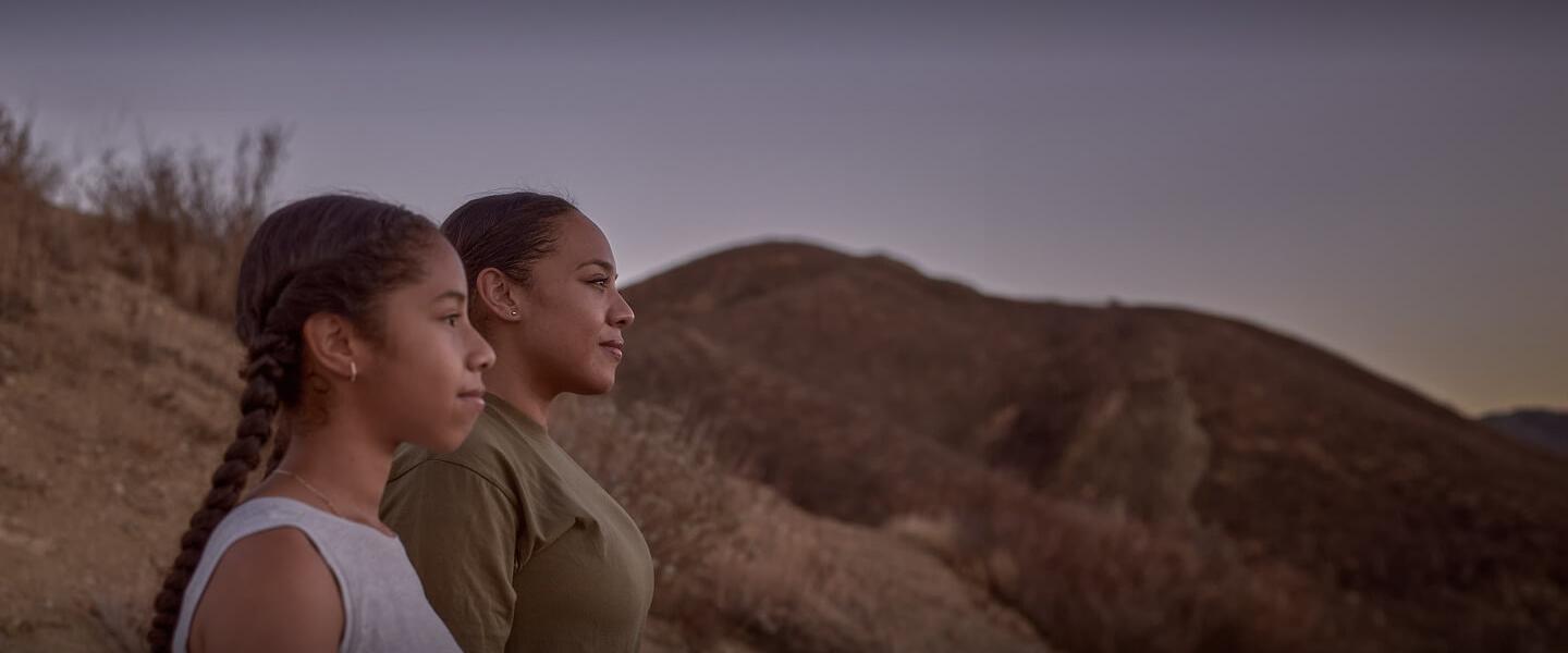 A mother and daughter stand together on a hill, silhouetted against a vibrant sunset.