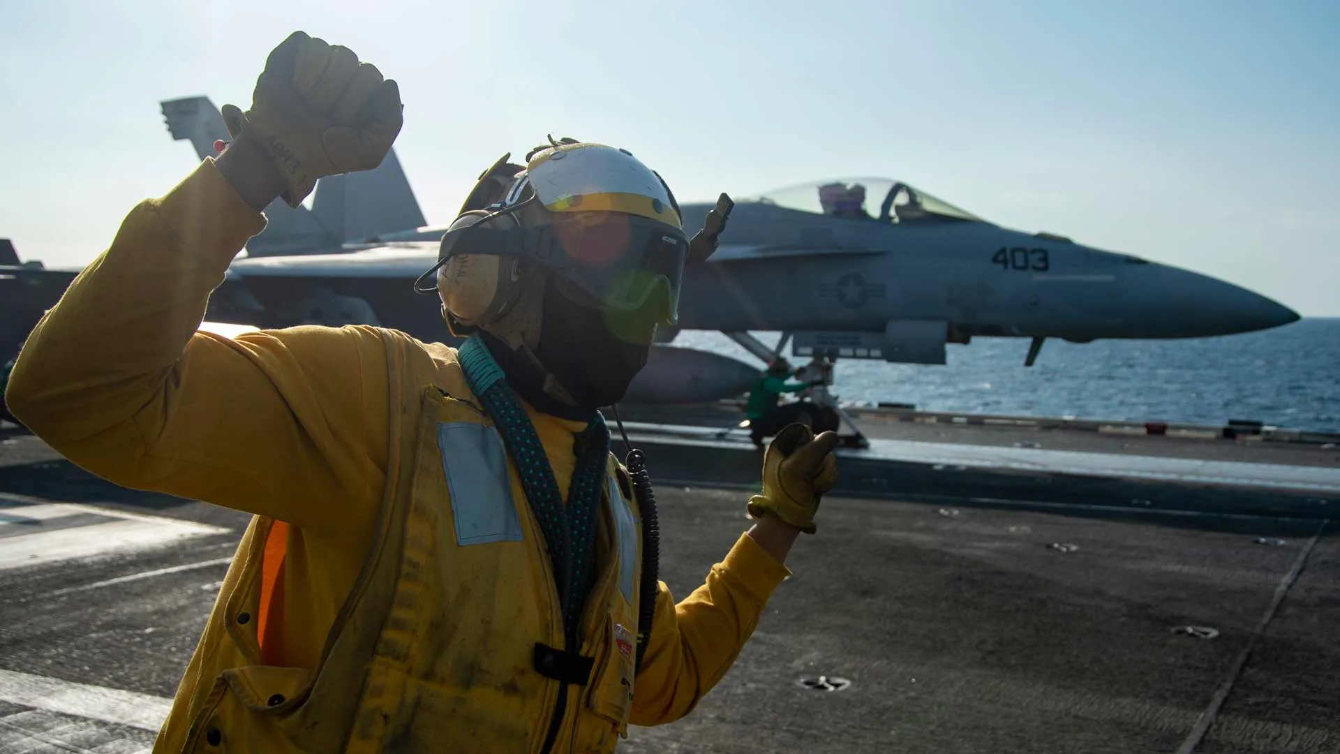 A United States Navy Aircrewman Operator gives hand signals to a Navy Pilot on the runway of an aircraft carrier